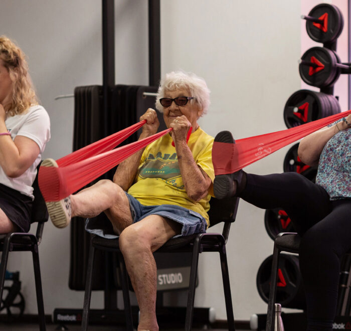 41be150d582f5927eb17fd57c54d2962e84bcb37 Three women in a gym setting in chairs using resistance bands