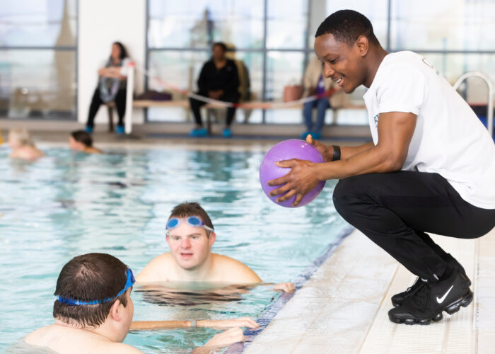 Our-Expertise-2 A man with a ball on the side of a swimming pool talking to two men in the pool