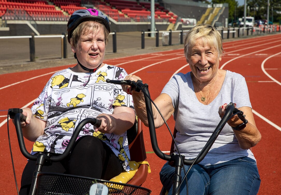 Two participants riding adapted cycles on an outdoor athletics track