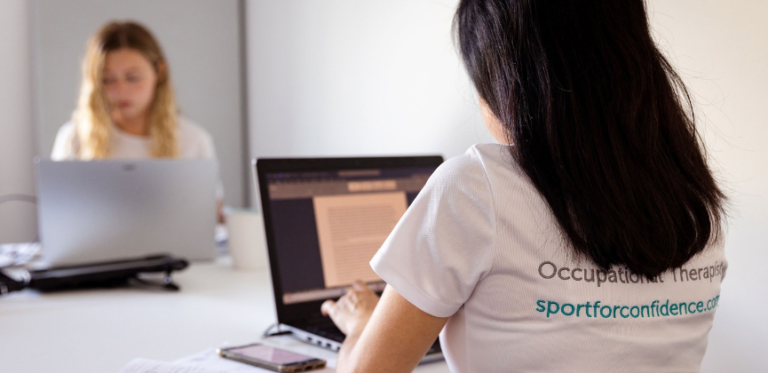 Woman wearing a “Sport for Confidence” occupational therapist shirt working on a laptop, with another person blurred in the background.