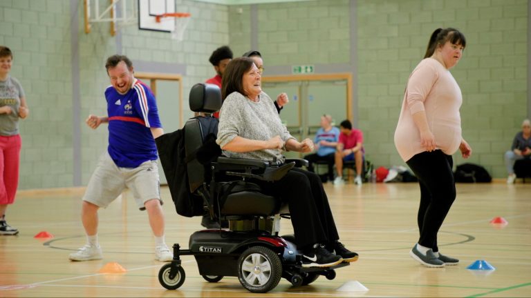 People of varying ages and abilities participate in an inclusive exercise session in a sports hall, smiling, moving, and engaging together.