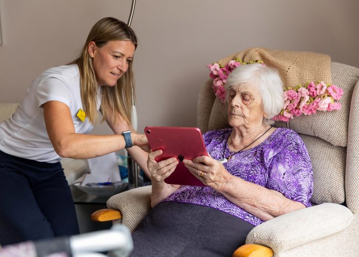 An occupational therapist supports an older woman to use an iPad who is sitting in an arm chair in her home.
