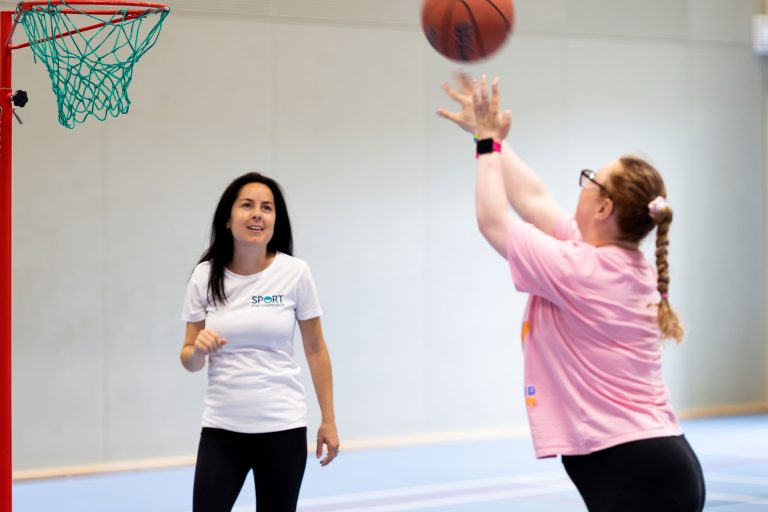 A Sport for Confidence occupational therapist looks on as a participant throws a basketball towards a hoop.