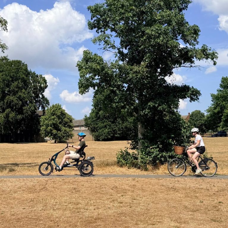 Two people cycling in an outdoor setting. The cyclist in front is riding an adaptive bicycle.